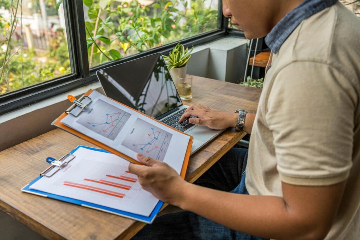 Person reviewing printed charts and graphs at a wooden desk while working on a laptop, with plants and windows visible in the background.