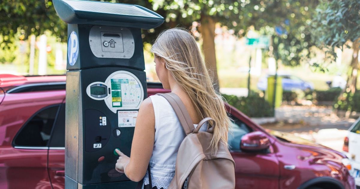 Woman using a public EV charging station in a parking lot surrounded by parked cars and trees.