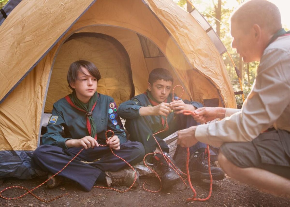 Two Boy Scouts in uniform learning rope knots from an adult instructor outside a tan camping tent in a wooded area.