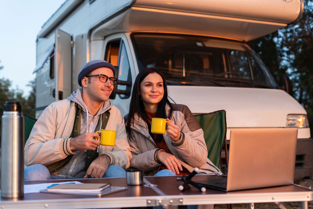 Couple holding yellow mugs sitting outside RV camper at dusk with laptop and thermos on table, looking at campground scenery.