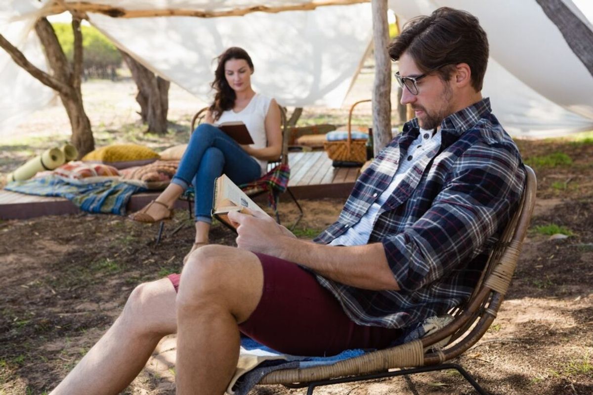 Man reading map in camping chair while woman holds book under tent canopy at outdoor glamping site.