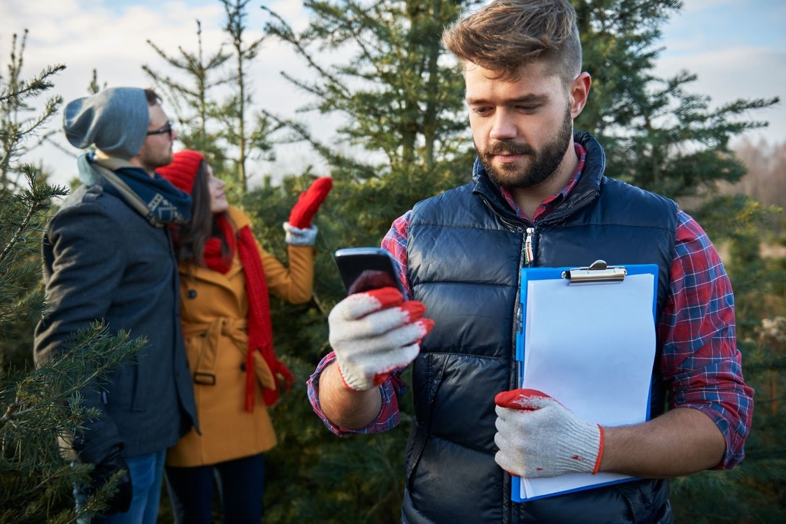 Man with clipboard and mobile device checking in at Christmas tree farm while couple shops in background.