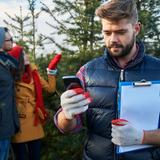 Man with clipboard and mobile device checking in at Christmas tree farm while couple shops in background.