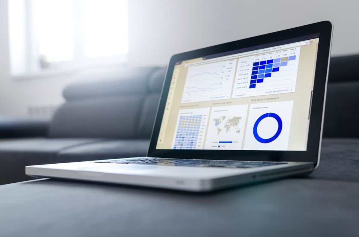 Laptop displaying business analytics dashboard with colorful charts, graphs, and data visualizations including a blue circular progress indicator and world map.