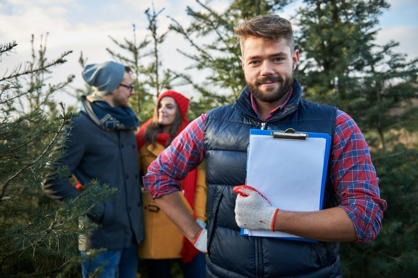 Bearded man holding clipboard in forest with three colleagues discussing campground operations among evergreen trees.