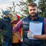 Bearded man holding clipboard in forest with three colleagues discussing campground operations among evergreen trees.