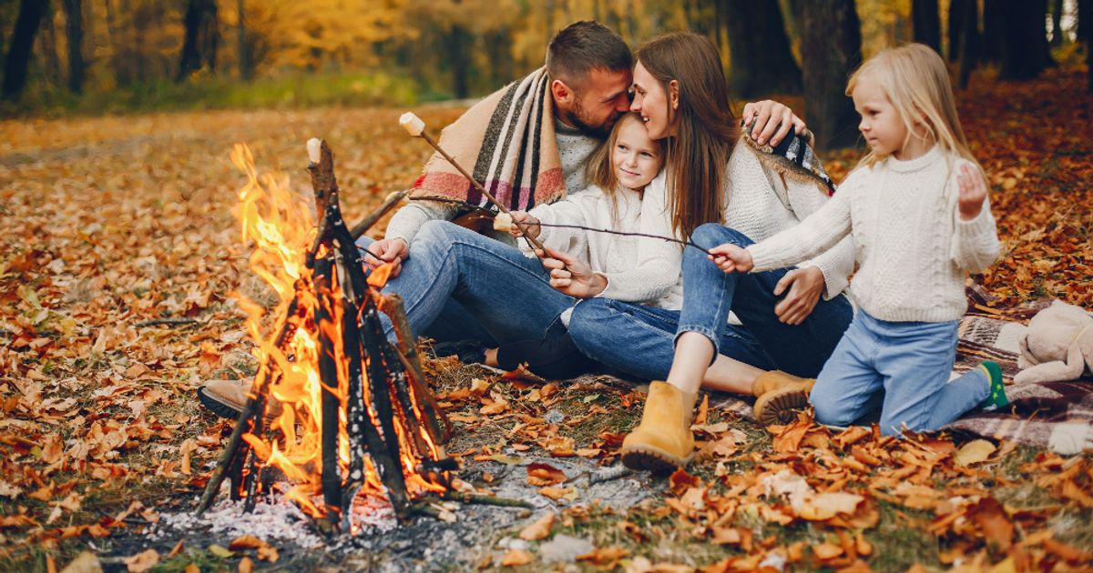 Family roasting marshmallows by a campfire in an autumn forest clearing surrounded by fall leaves.