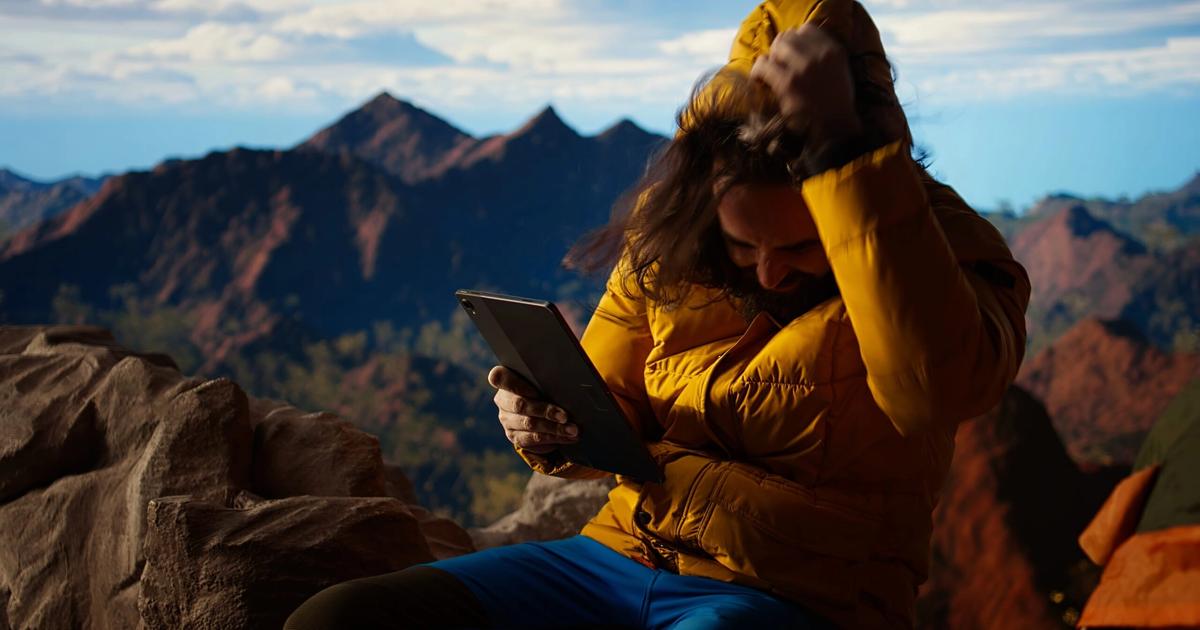 Woman in yellow jacket using tablet while sitting on rocks with mountain landscape in background