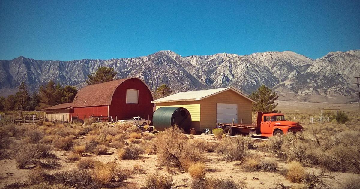 Rural farm property with red barn, tan building, orange vintage truck, water tank, and mountains in background under clear sky.
