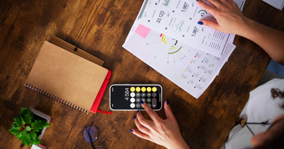 Person reviewing financial data on printed charts and spreadsheets at wooden desk while using calculator app on smartphone.