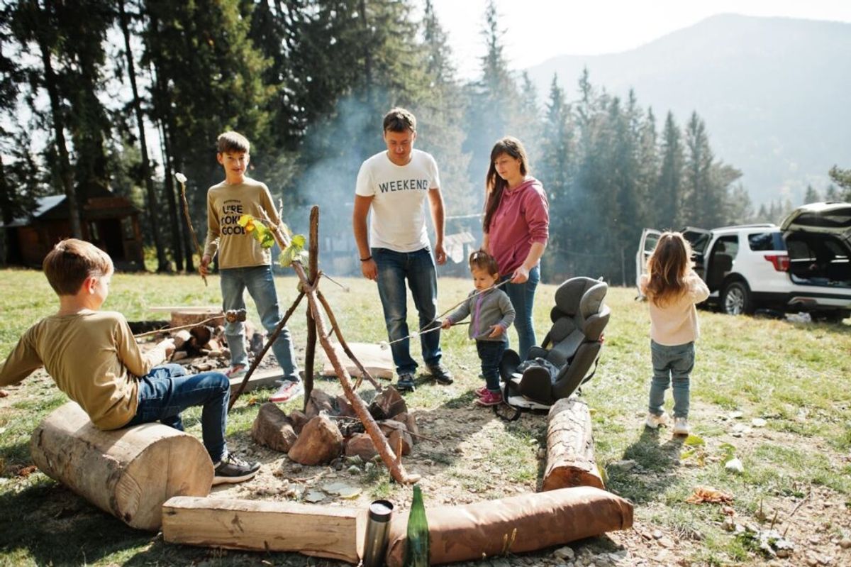 Family gathered around campfire at mountain campground with children roasting marshmallows and adults supervising in forest setting.