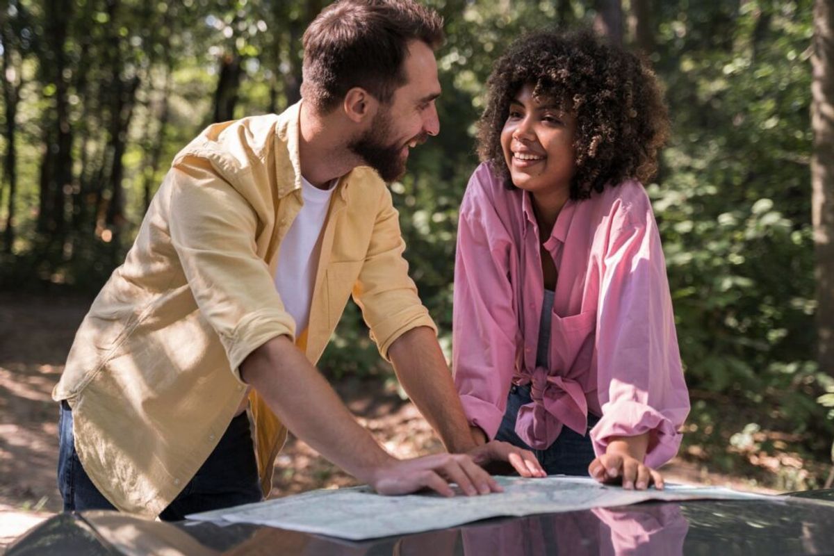 Two people reviewing a map together on a wooden table in a sunlit forest setting surrounded by trees.