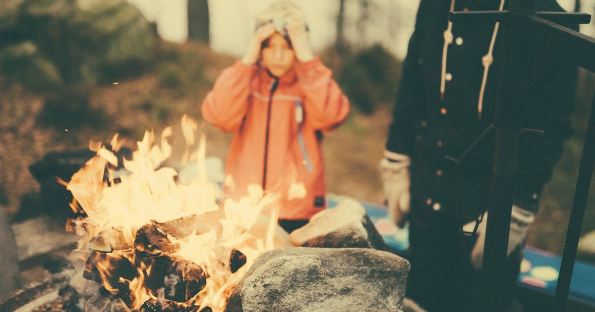 Family gathered around a campfire outdoors, with child in orange jacket and adult in dark coat enjoying the flames and warmth together in a forest setting.