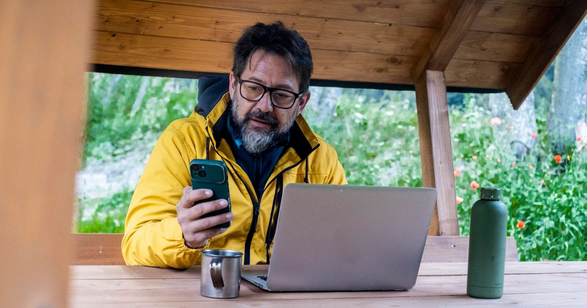Man in yellow jacket working on laptop at wooden desk in covered outdoor space, holding smartphone, with green water bottle and metal cup nearby, mountain landscape visible in background.