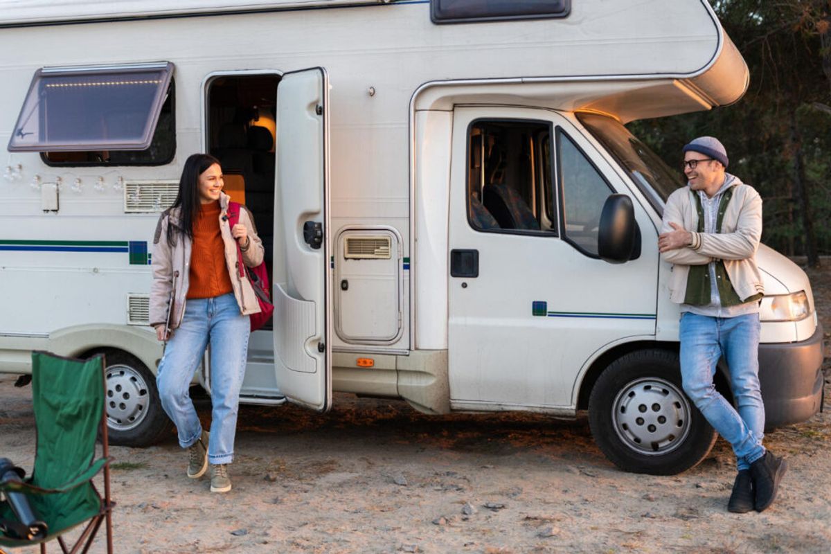 Couple standing outside white RV camper van at campground, smiling and conversing, with camping chair visible nearby.