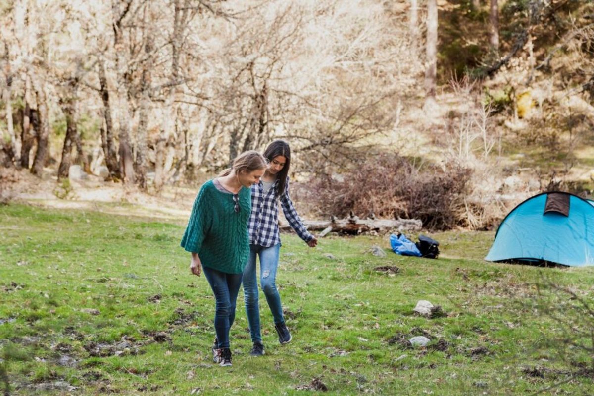 Two women walking together at a camping site with a blue tent and bare trees in the background on grassy ground.