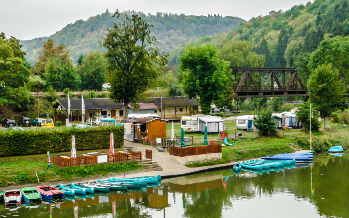 Riverside RV park with colorful kayaks moored in water, trailers parked on grass, wooden structures, trees, mountains, and a bridge in background.