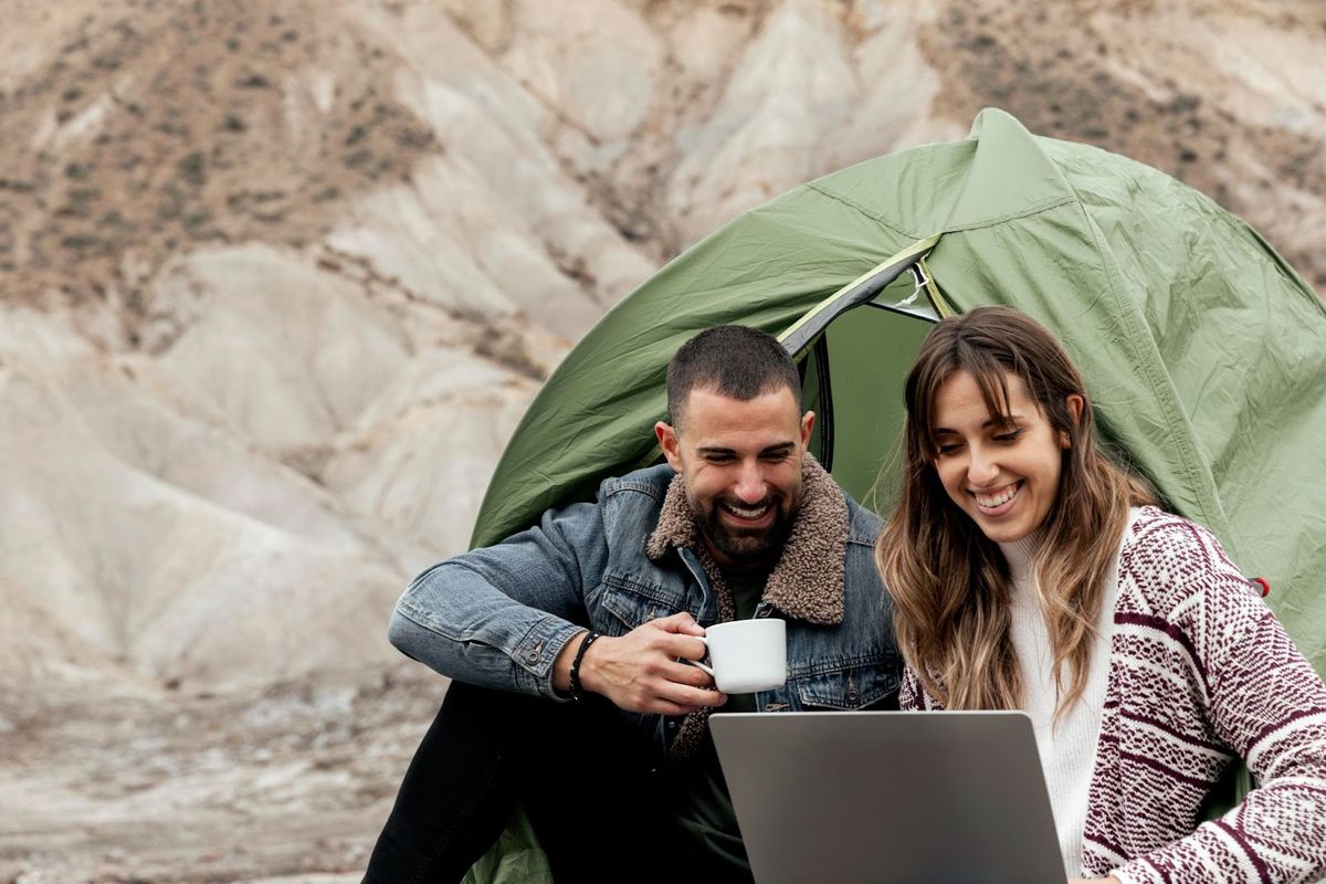 Couple under green tent reviewing laptop while holding coffee mug in rocky mountain landscape setting.