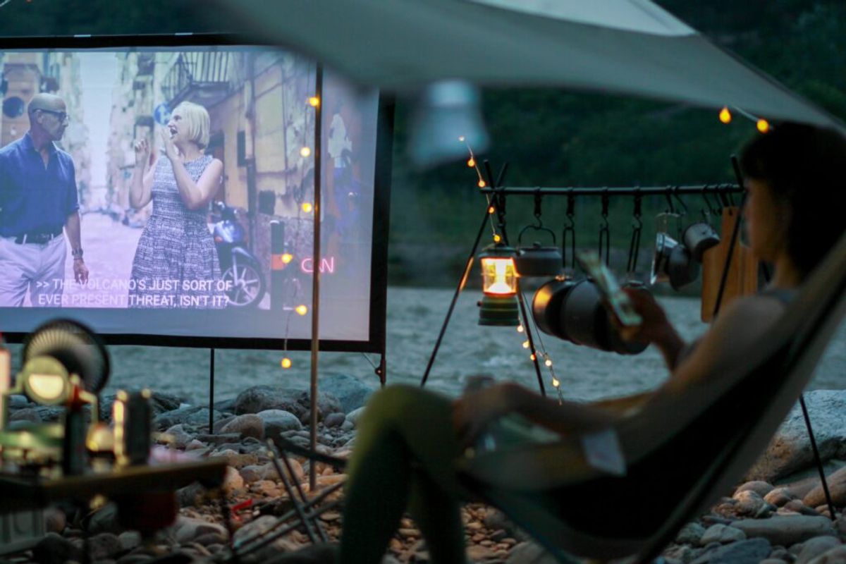 Outdoor movie night at a glamping site with guests watching a film on a projected screen, surrounded by string lights and camping equipment on a beach setting.