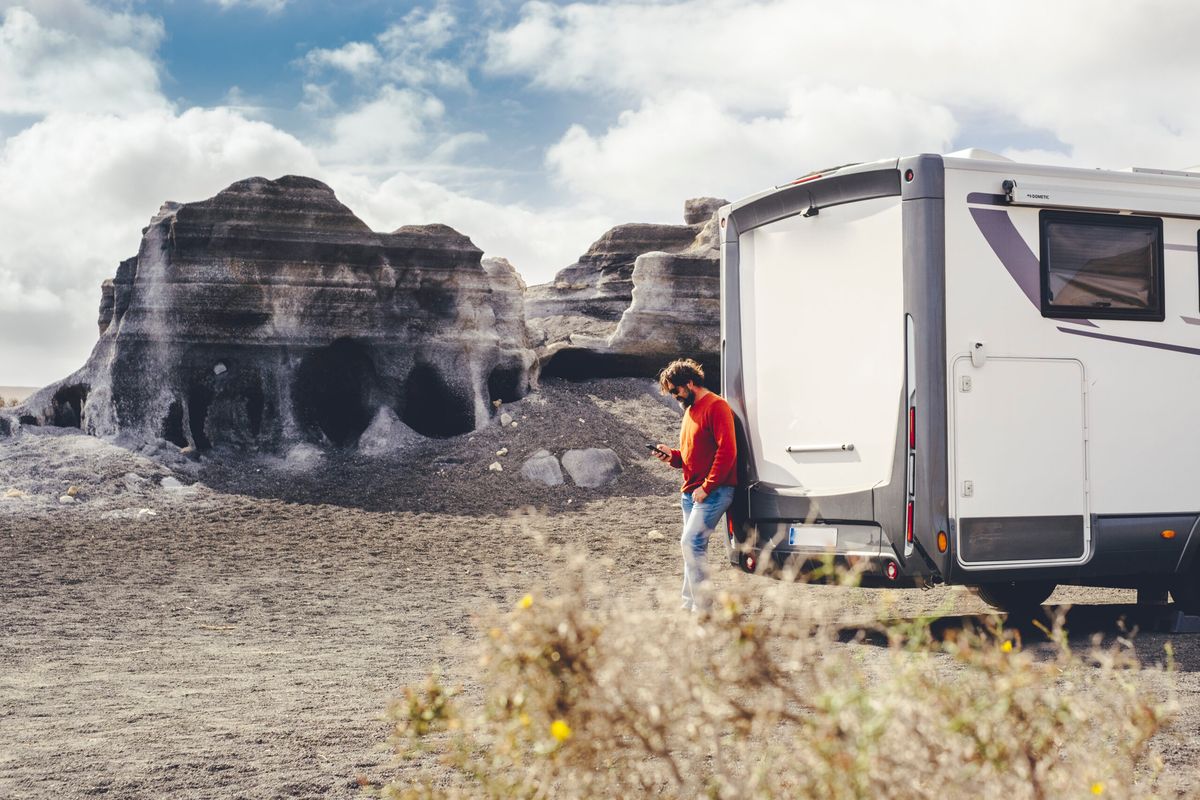 Person in red jacket standing by white RV camper parked near ancient rock formations in desert landscape under cloudy sky.