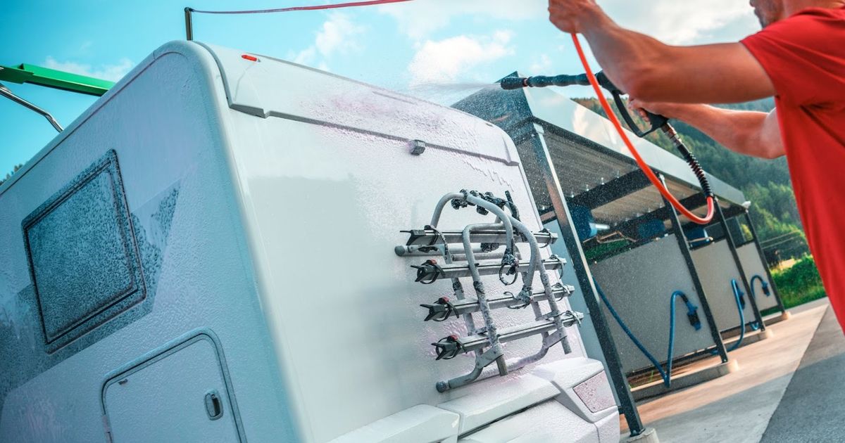 Man in red shirt connecting water hose to back of white RV with bike rack mounted on rear, mountains visible in background.