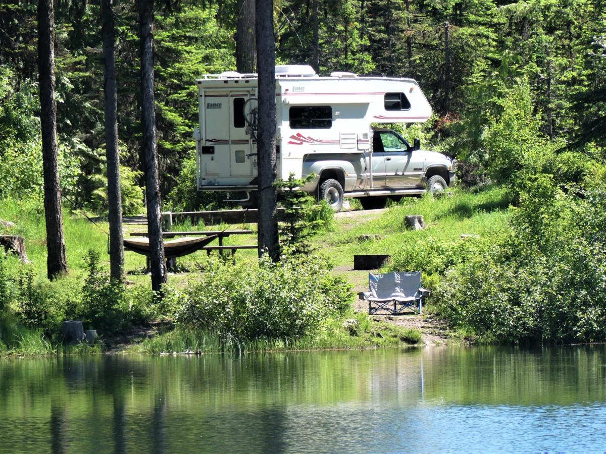 Pickup truck camper parked at a wooded campsite beside a calm lake, with picnic table and camping chair visible among trees.