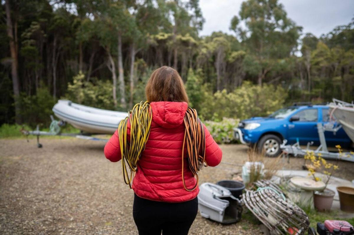 Woman in red jacket carrying coiled yellow rope at campground with boat, blue truck, and forest in background.
