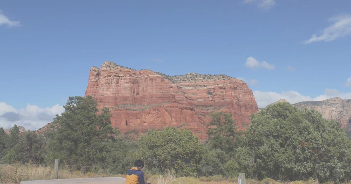 Person sitting at picnic table overlooking red rock formation and pine forest under blue sky with clouds.