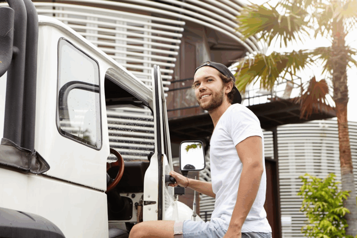 Man in white shirt opening door of white RV parked at residential campground with palm trees and modern pergola structure visible in background.