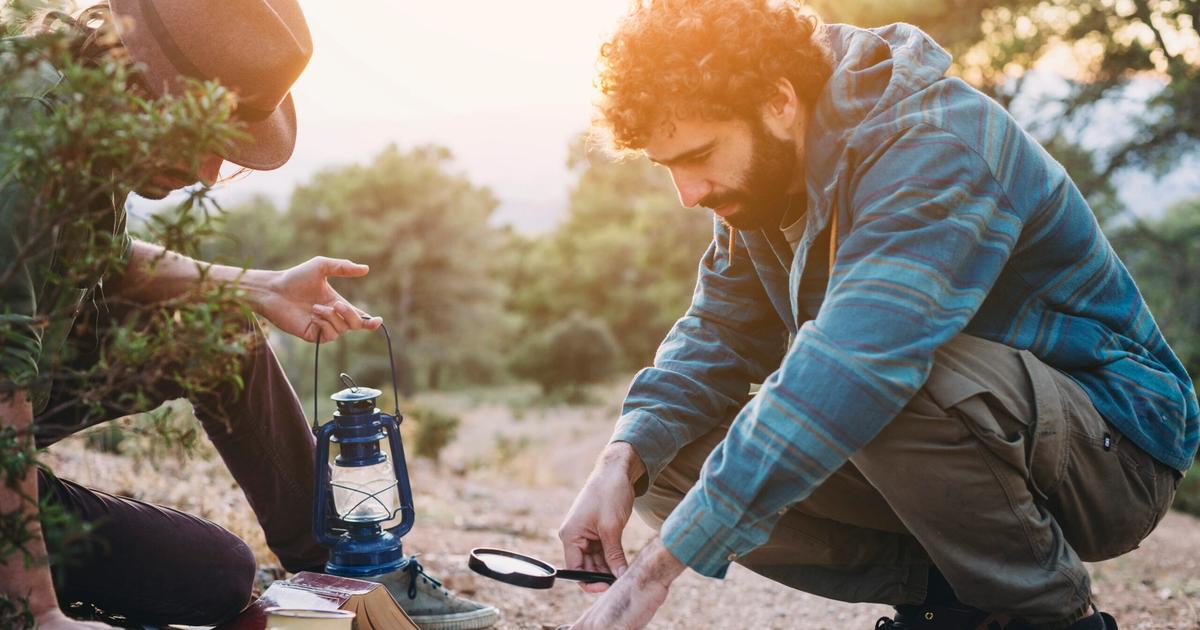 Two campers review a map together outdoors at dusk, with a lantern and coffee mug on the ground beside them.