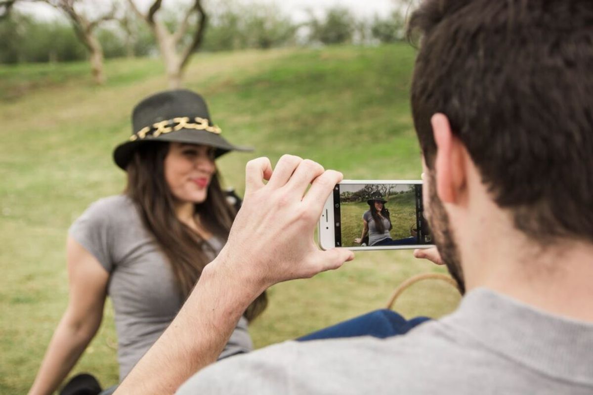 Man taking a photo of woman in black hat at outdoor campsite with green field and trees in background