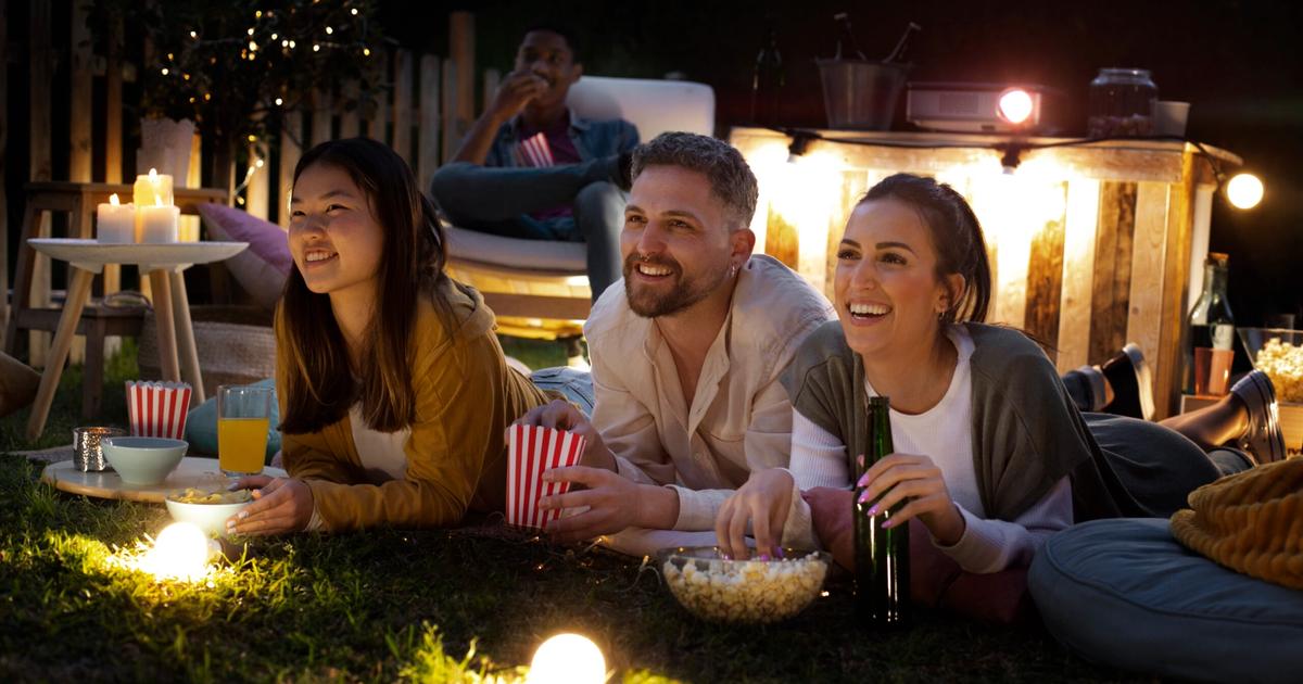 Group of three friends enjoying outdoor movie night at RV park, lying on grass with popcorn and drinks while watching film projected on screen.