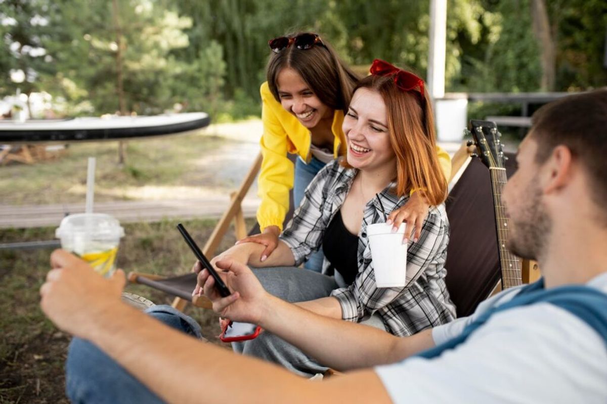 Group of young adults laughing together outdoors at campsite, holding drinks and a guitar, enjoying recreational activities.