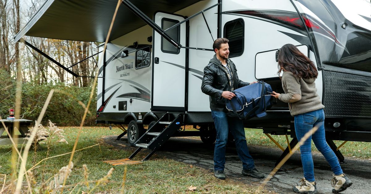 Couple outside a North Trail travel trailer at a campsite, man holding a blue bag while woman gestures toward the RV's entrance.