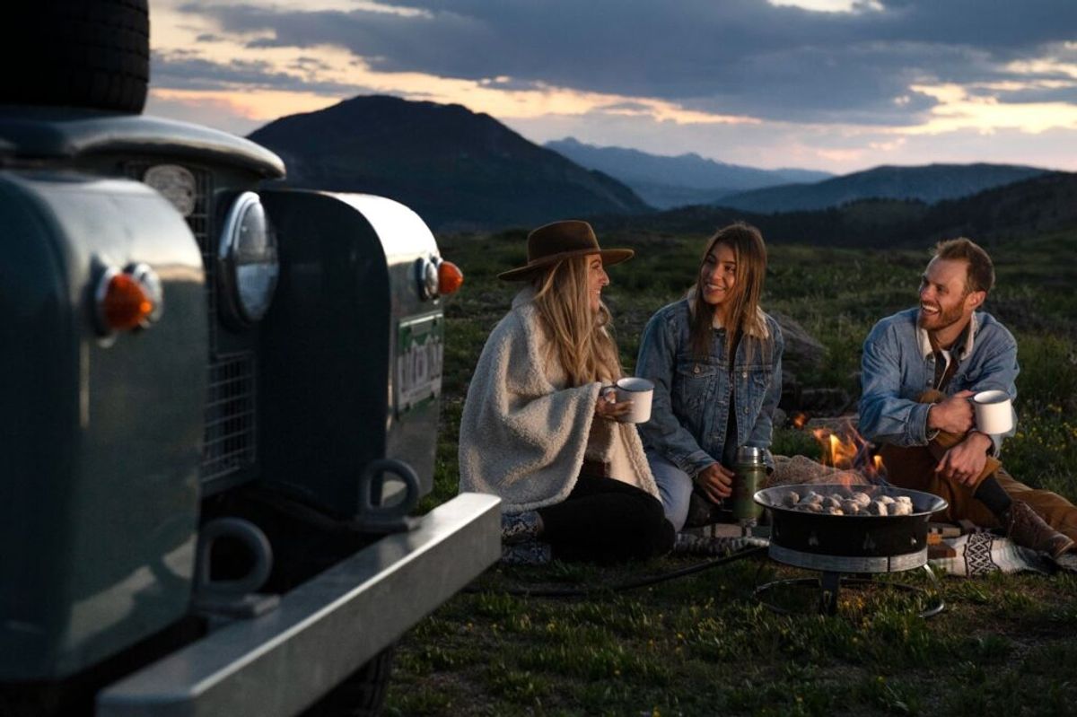 Three friends enjoy campfire cooking and socializing near a vintage Airstream trailer with mountains in the background at dusk.