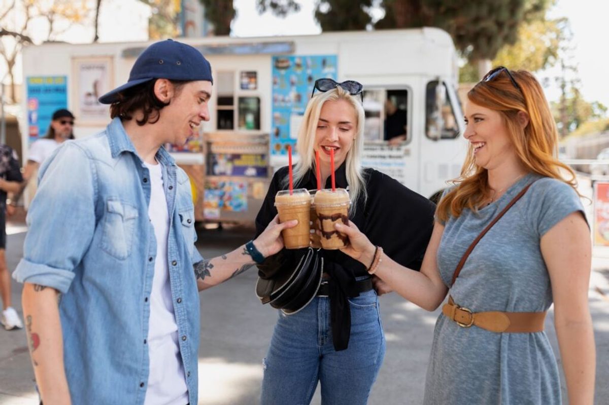 Three young people cheering with iced beverages in front of a food truck at an outdoor event.