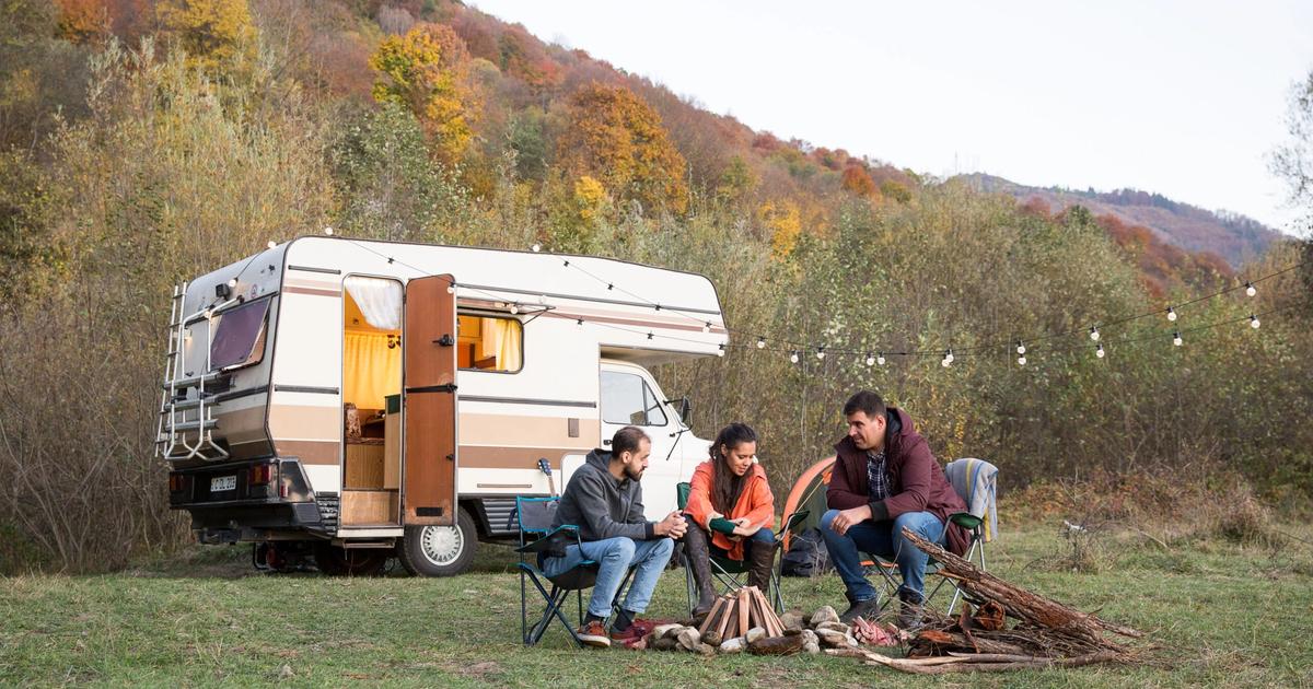 Four people sitting around a campfire next to a white RV camper in an autumn forest setting.