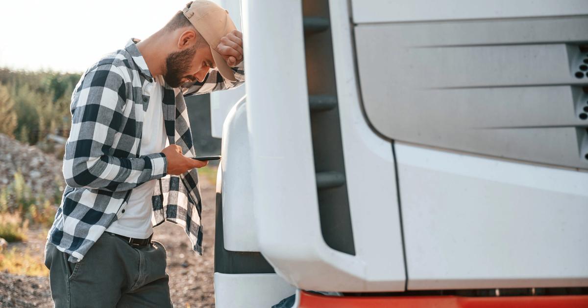 Man in plaid shirt and cap inspecting RV exterior door, holding clipboard, with forest and rocky terrain visible in background.