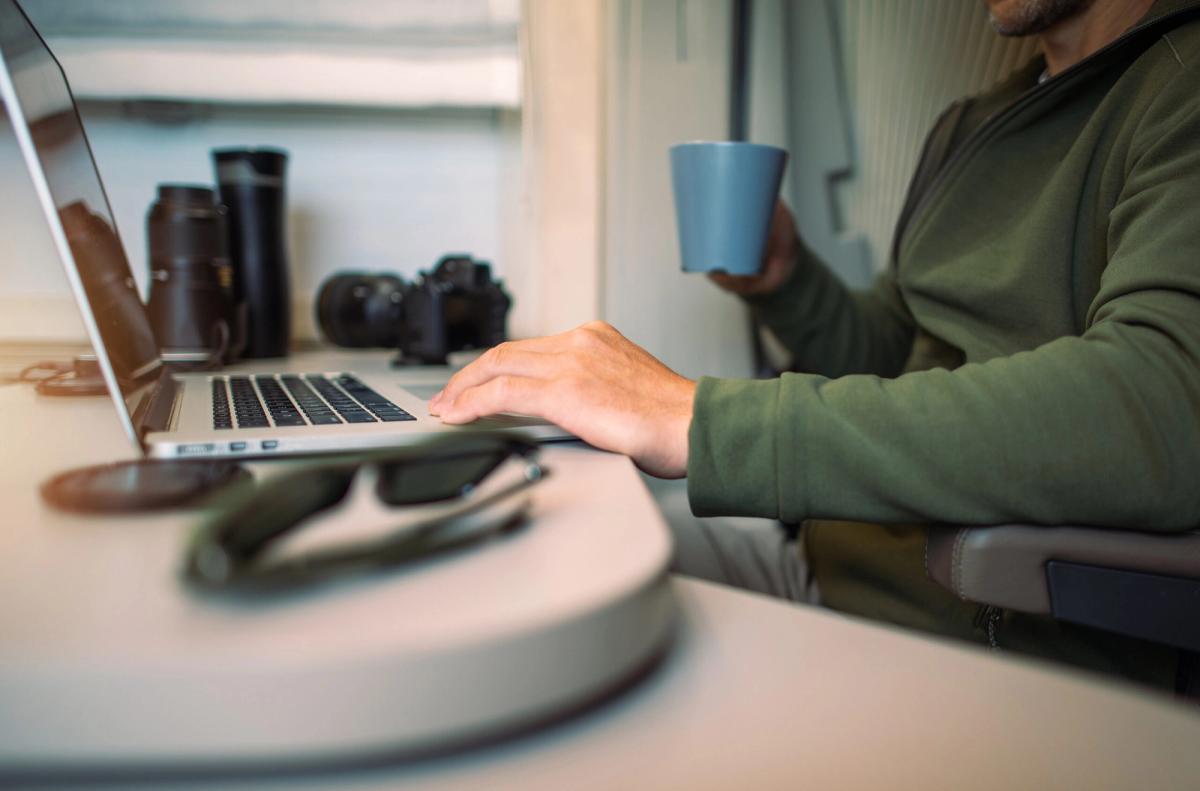 Person in olive jacket working on laptop at desk with camera equipment and coffee cup nearby, suggesting remote work setup at campground facility.