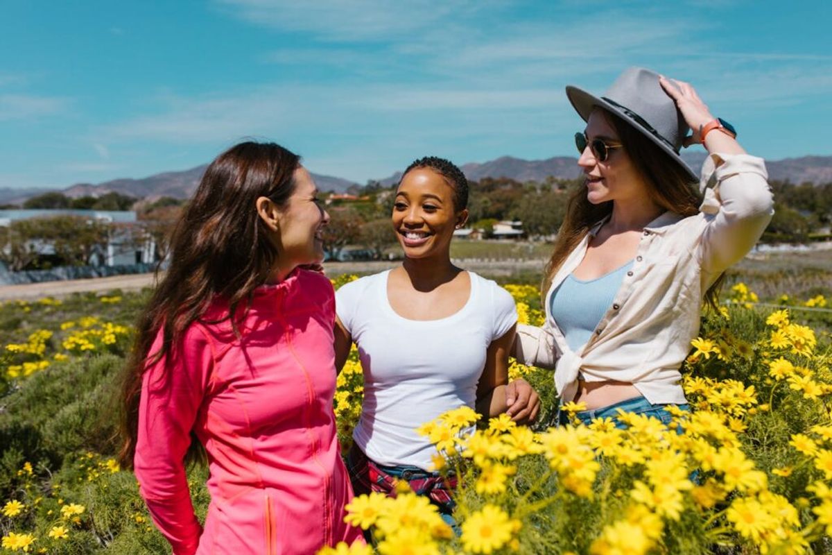 Three women smiling in a yellow wildflower field with mountains and water visible in the background under blue sky.