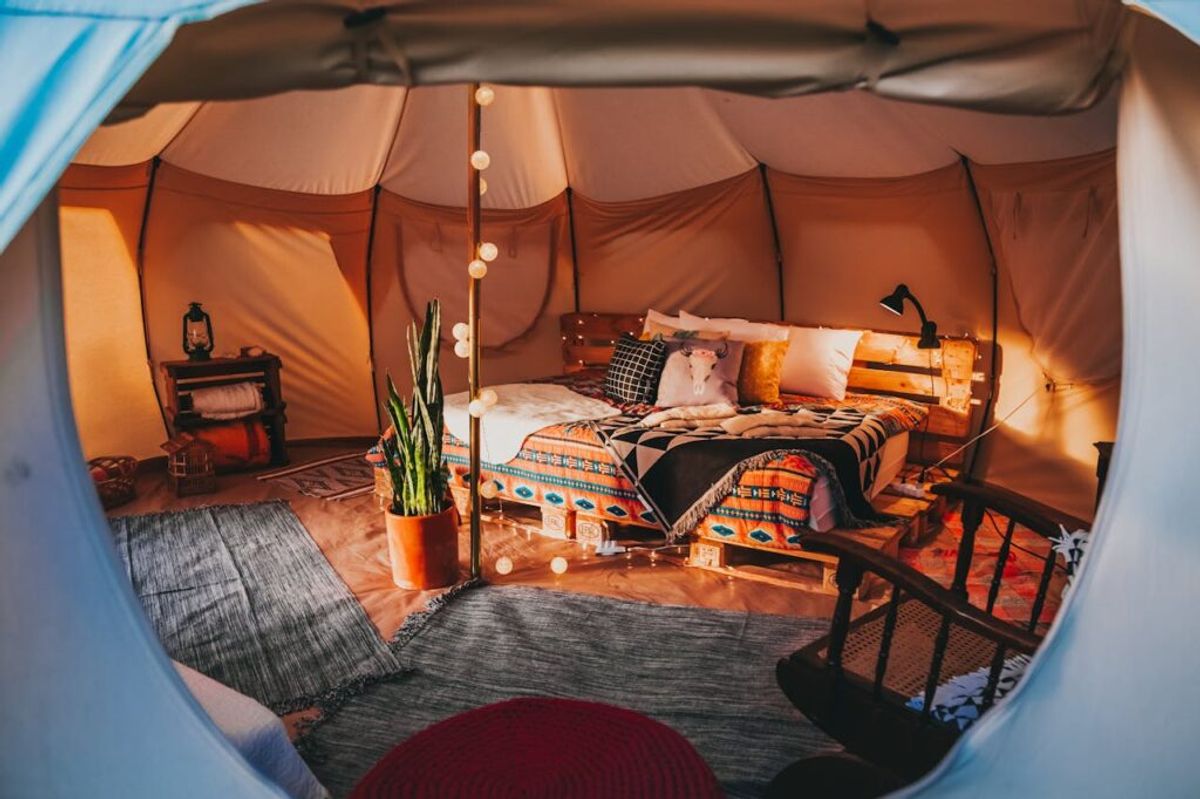 Luxurious glamping tent interior featuring an orange fabric structure, bed with patterned bedding, potted plant, string lights, and wooden furniture.