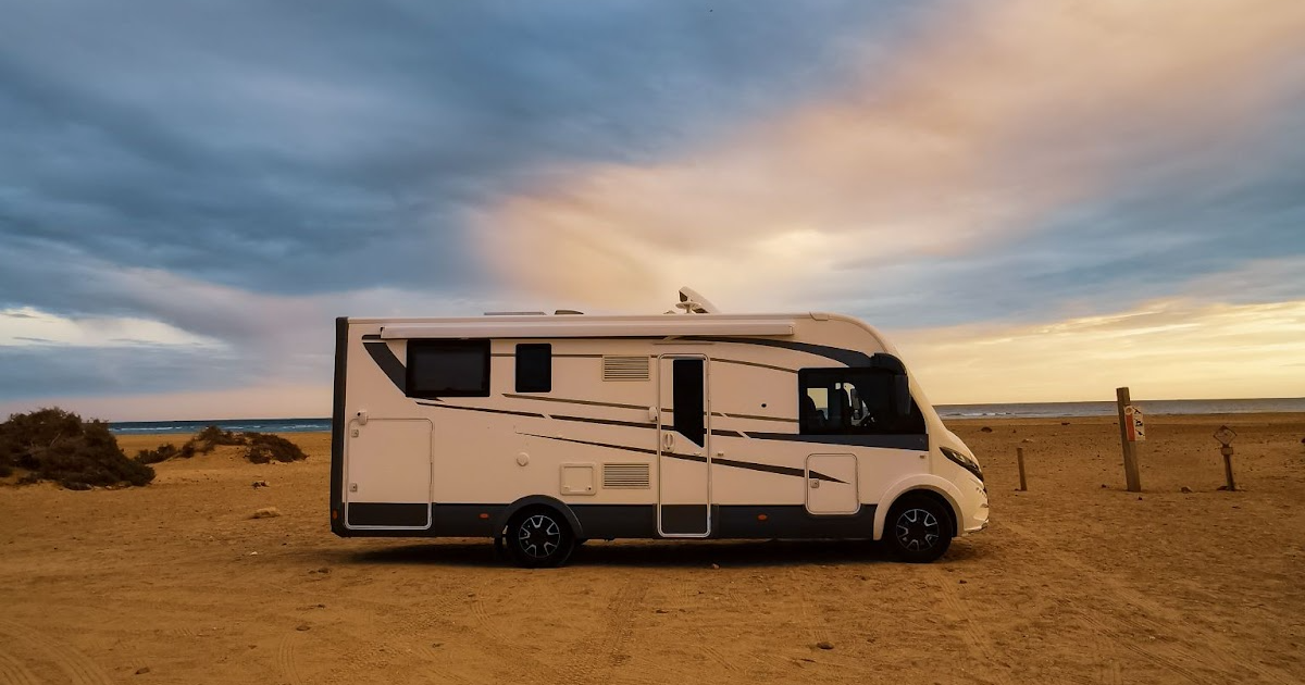 Beige motorhome parked on sandy desert terrain with ocean and dramatic cloudy sky in background, fence line visible in distance.