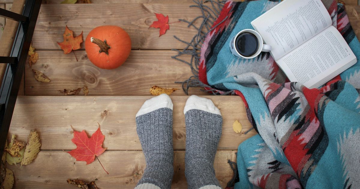 Person wearing cozy socks relaxing on a blanket with an open book, coffee mug, pumpkin, and autumn leaves on a wooden floor.