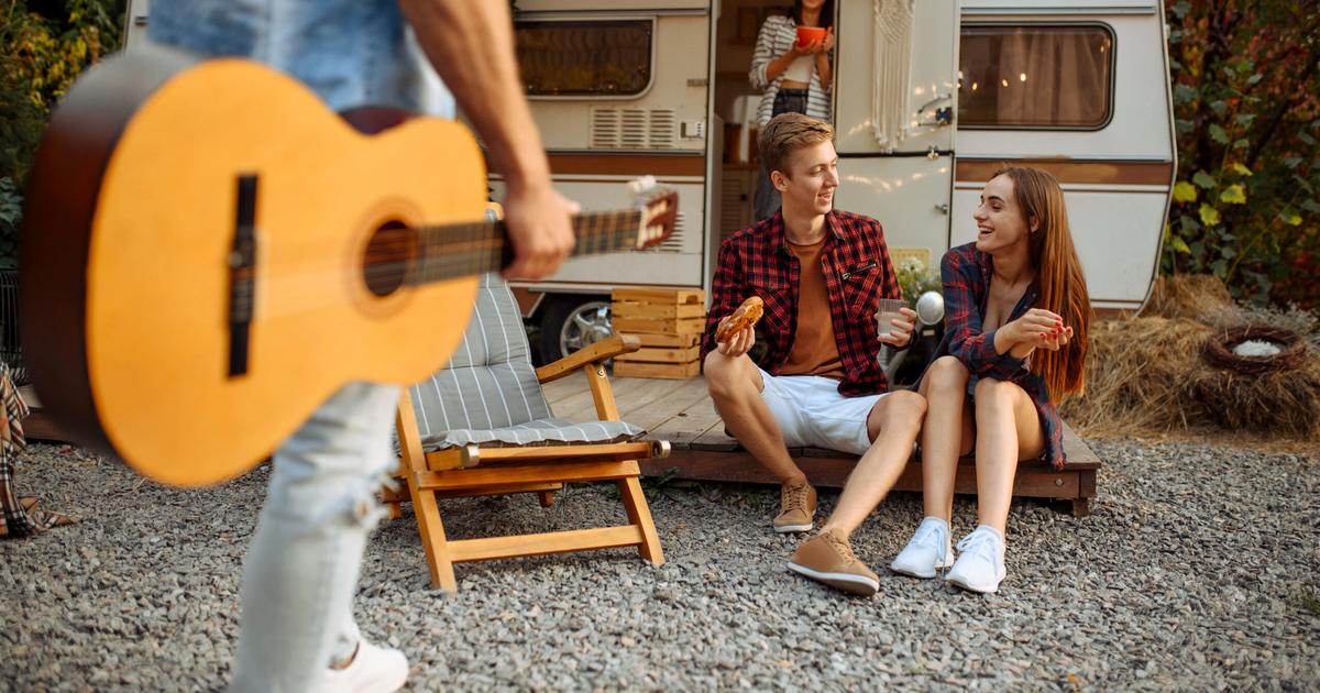 Young adults enjoying campfire gathering outside RV trailer, one playing acoustic guitar while others sit on chairs eating and socializing.