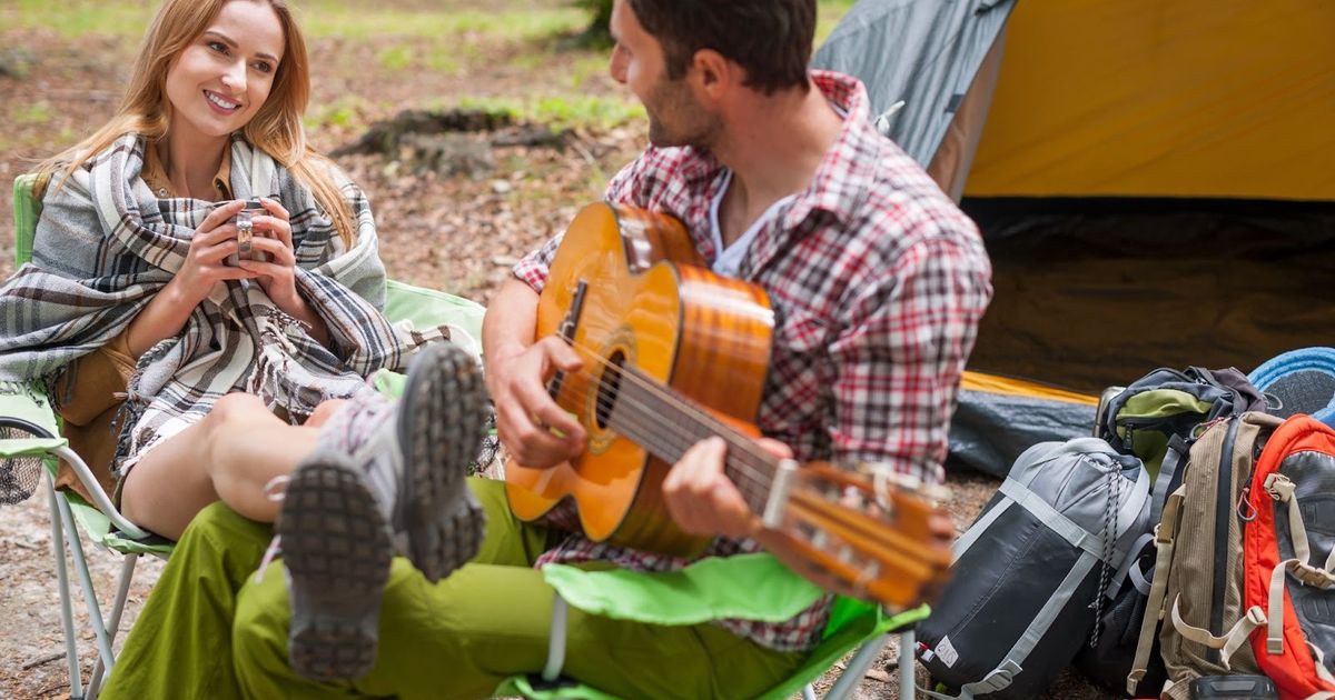 Couple camping by tent with man playing acoustic guitar while woman holds warm drink and smiles, surrounded by backpacks and camping gear.