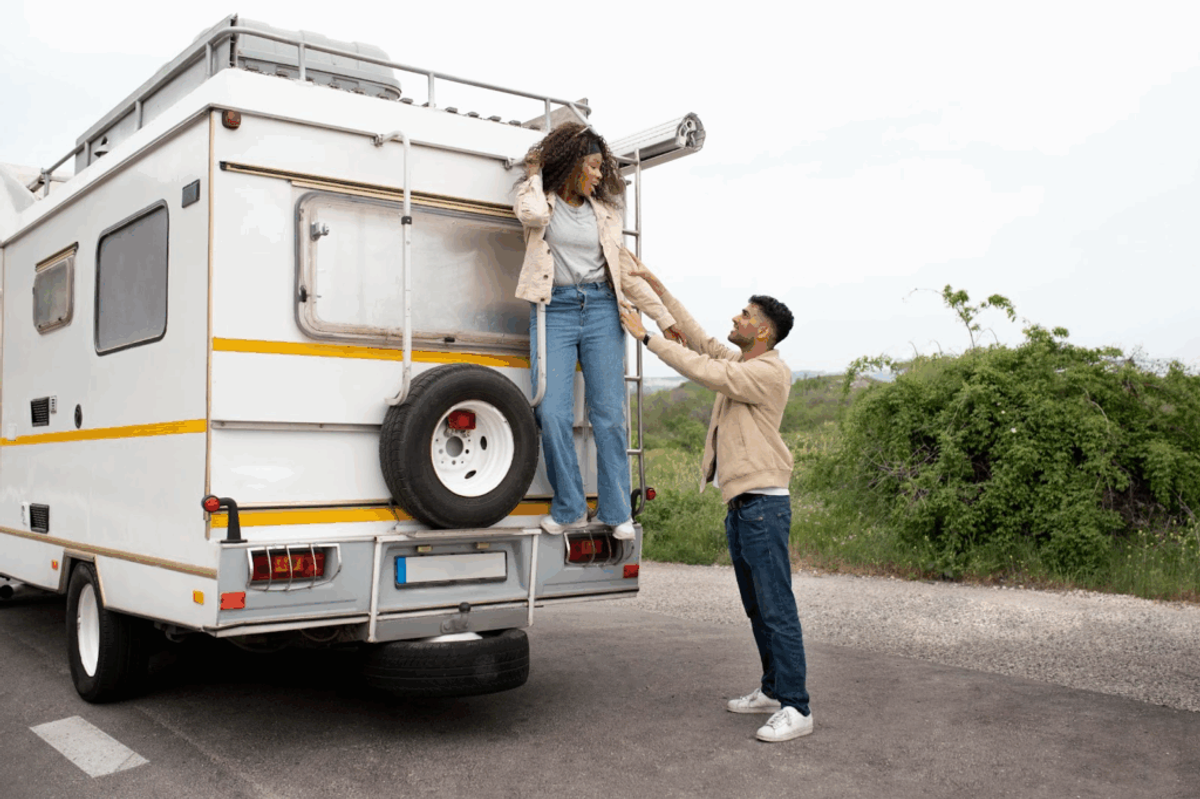 Couple at RV campsite, man helping woman climb onto roof of white and yellow motorhome with green landscape in background