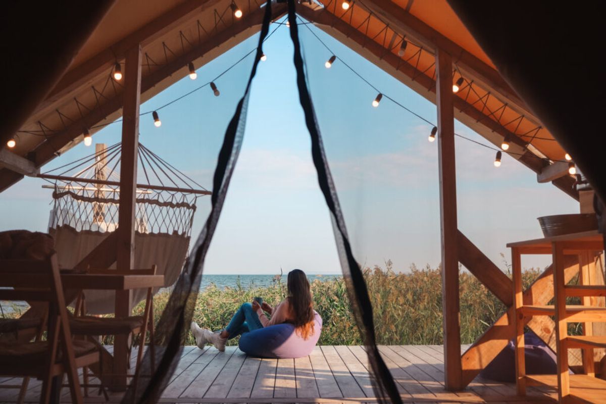 Person relaxing in hammock on wooden deck overlooking ocean and trees, with pergola and hanging lights in glamping setup.