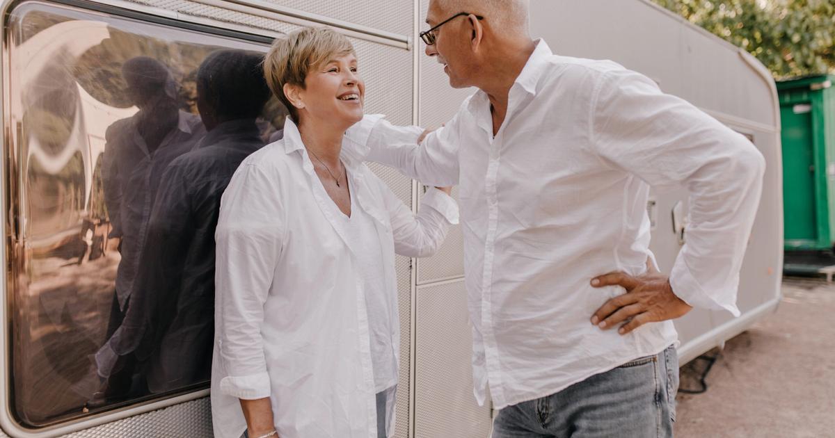 Senior couple in white shirts smiling while boarding an RV at a campground with ivy-covered structures visible in background.