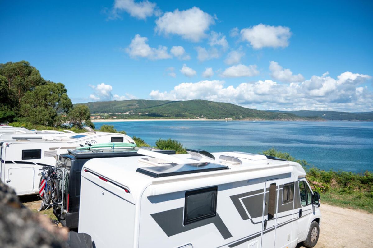 Motorhomes parked at a coastal campsite overlooking a blue ocean bay with green hills and clear sky.