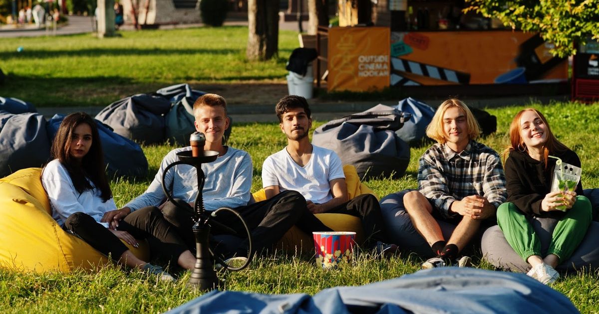 Group of five young people sitting on bean bags in a grassy park area, holding drinks and snacks during a casual outdoor gathering.